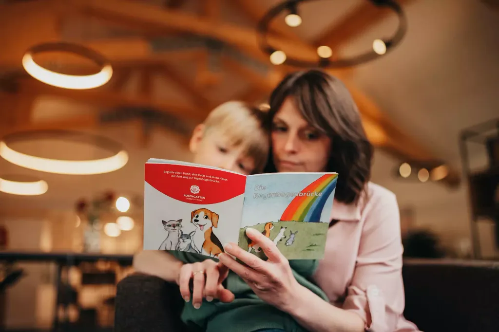 Kobieta i dziecko wspólnie czytają książkę dla dzieci „Tęczowy most” z ilustracjami przedstawiającymi psa, kota i zająca.