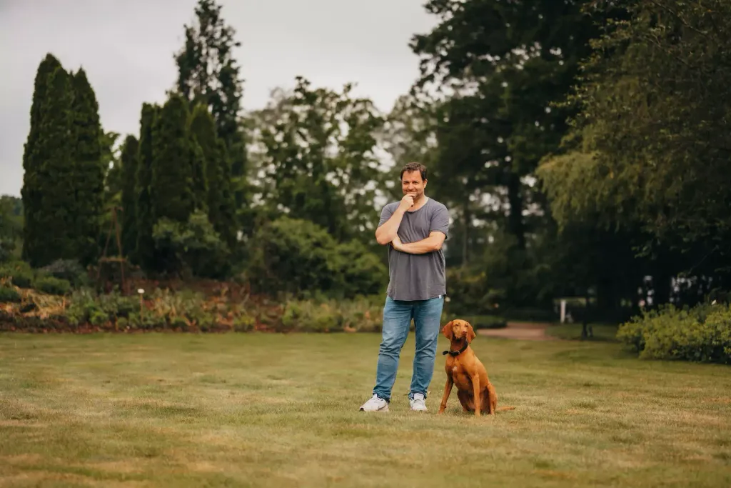 Martin Rütter stoi na łące w parku. Obok niego siedzi pies.